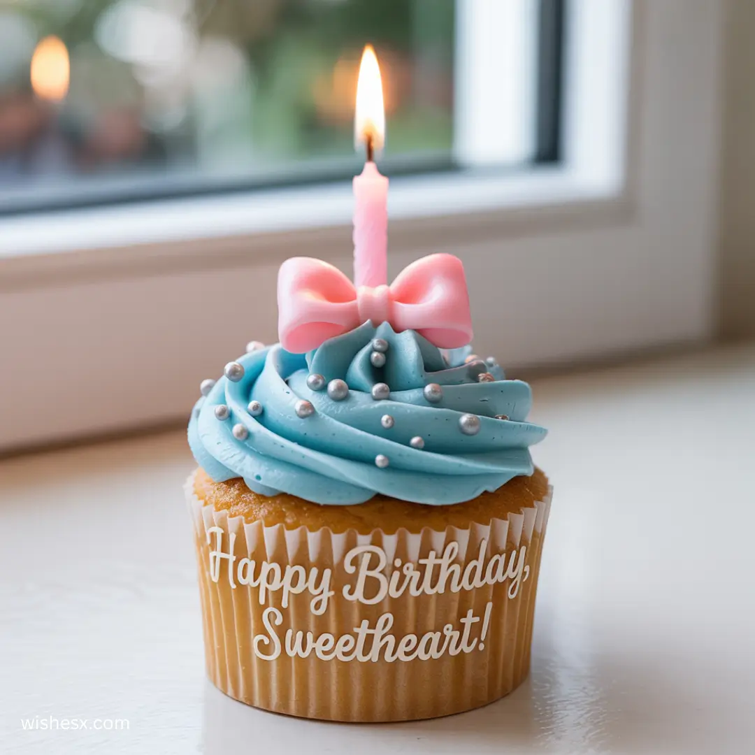 A cupcake with blue frosting, silver pearls, and a pink bow, topped with a lit candle. The wrapper reads "Happy Birthday, Sweetheart!" in white script.